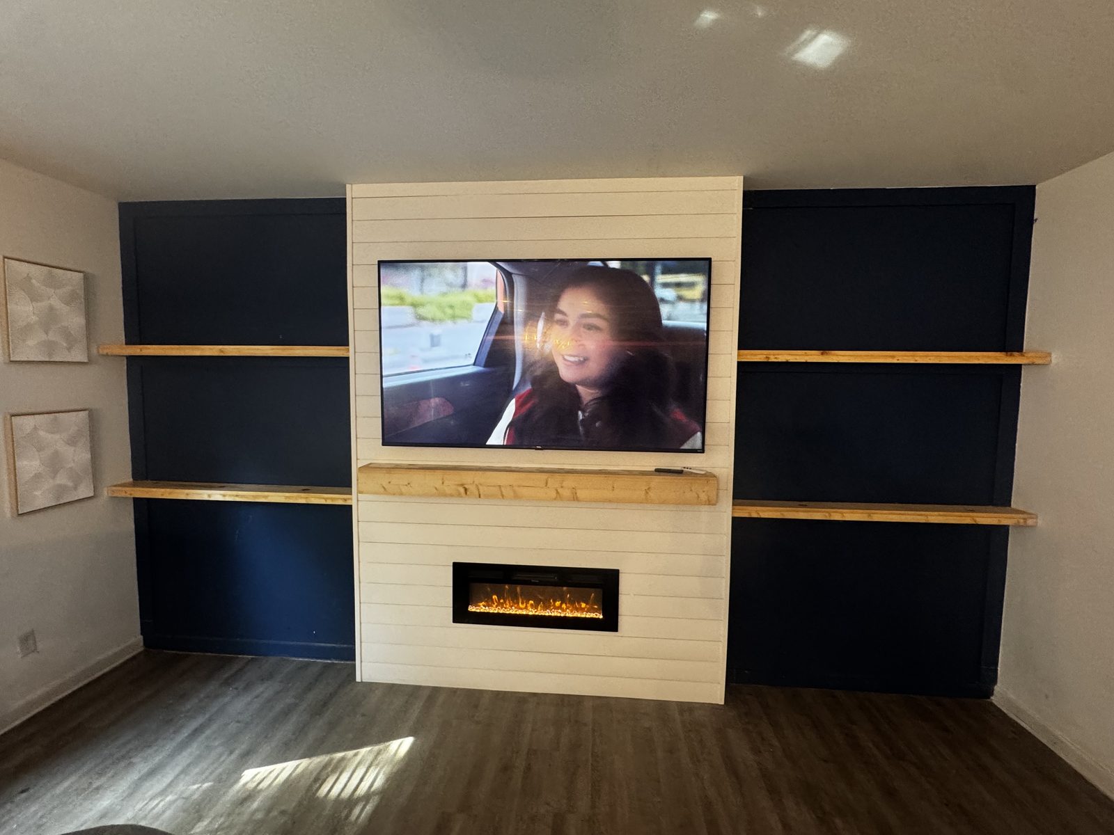 TV mounted on a shiplap wall over an electric fireplace, with floating wood shelves on both sides